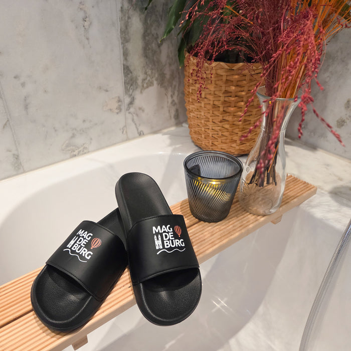 Schwarze Magdeburg Sandalen auf einer Badewannenablage, begleitet von dekorativen Elementen wie einem Kerzenhalter und getrockneten Blumen in einer Vase. MagdeLetten Design im Fokus.