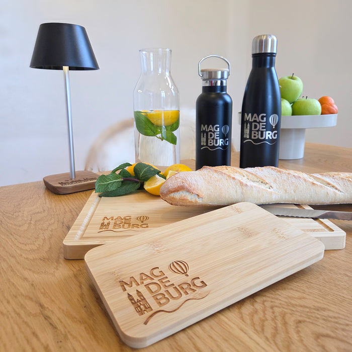 Holztisch mit Magdeburg-Themenartikeln: zwei schwarze Edelstahlflaschen mit 'Magdeburg' Schriftzug, Glaswasserkaraffe mit Zitronen und Minze, zwei Schneidebretter mit Magdeburg-Gravur und ein Baguette. Im Hintergrund eine Schale mit Obst. Perfekt für Küche und Picknick.