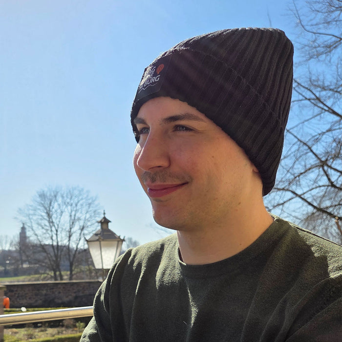 Junger Mann mit schwarzer MagdeBeanie-Mütze im Freien, Hintergrund zeigt einen klaren Himmel und Bäume, städtische Szene in Magdeburg hervorgehoben.