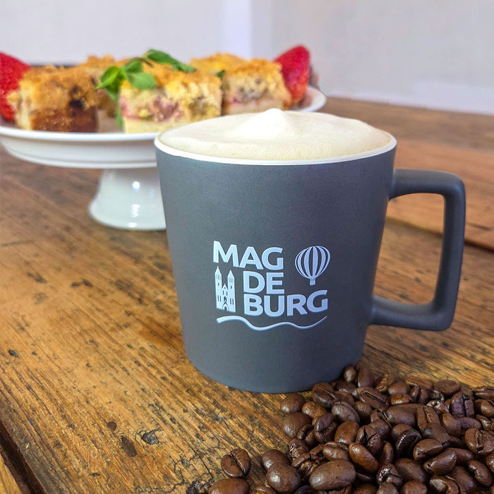 Graue Tasse mit Magdeburg-Logo und Kaffee-Schaum gefüllt, daneben Kaffeebohnen auf einem Holztisch, im Hintergrund ein Teller mit dekorativen Kuchenstücken.