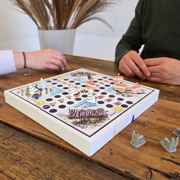 Zwei Personen spielen das Magdeburger Würfelspiel auf einem Holztisch. Das Spielbrett zeigt bunte Spielfiguren und historische Gebäude von Magdeburg. Im Hintergrund sind dekorative Gräser zu sehen. Brettspiel, Magdeburg, Würfelspiel.