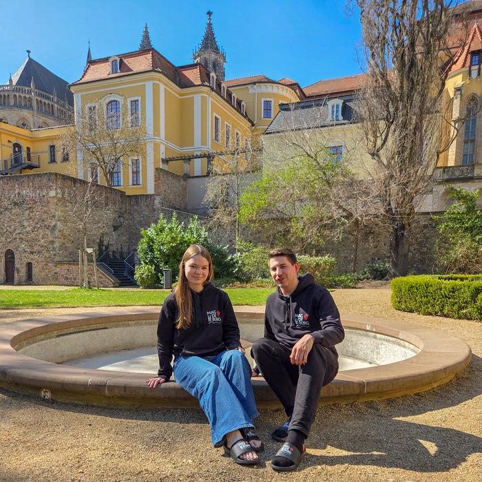 Zwei Personen sitzen auf einem Brunnenrand in einem sonnigen Garten. Beide tragen den stilvollen Unisex-Hoodie im Magdeburg-Look. Im Hintergrund ist ein historisches Gebäude mit gelber Fassade und roten Dächern zu sehen. Diese Szenerie spiegelt den stilvollen und urbanen Charakter von MagdeHoodie wider.