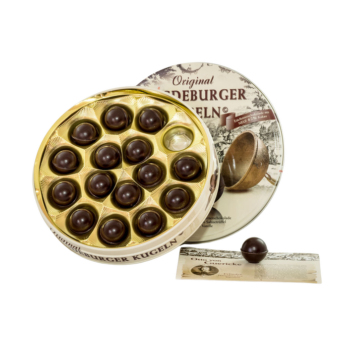Eine geöffnete Schachtel Magdeburger Kugeln Edelbitter Pralinen, gefüllt mit dunklen Schokoladenkugeln in goldener Verpackung, daneben der Deckel mit historischer Illustration und eine Kugel auf einem Flyer.