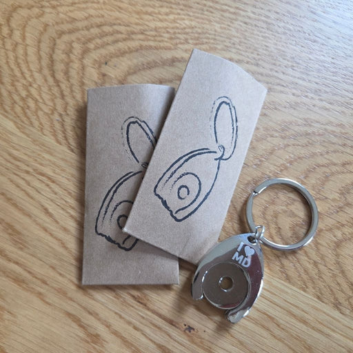 Das Bild zeigt zwei braune Papierhüllen mit einer schwarzen Illustration eines Einkaufswagenchips im Schlüsselanhänger-Design. Daneben liegt ein silberner Schlüsselanhänger in Form eines Einkaufswagenchips mit der Gravur „I ❤️ MD“ (Ich liebe Magdeburg) auf einer Holzoberfläche.