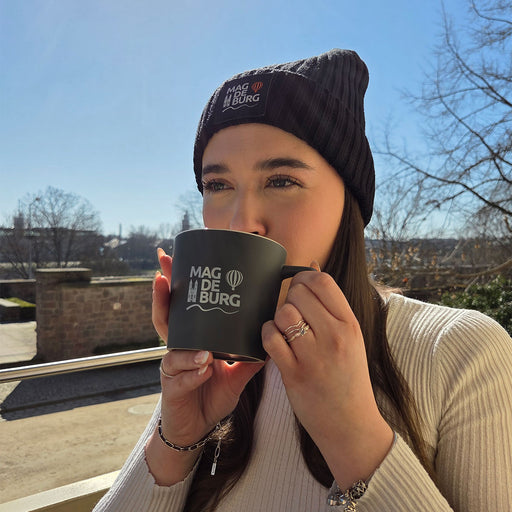 Frau mit schwarzer Mütze und schwarzer Tasse mit Magdeburg-Logo sitzt im Freien und genießt das sonnige Wetter, hinter ihr Skyline von Magdeburg sichtbar. Outdoor-Szene, Fokus auf regionalem Design.