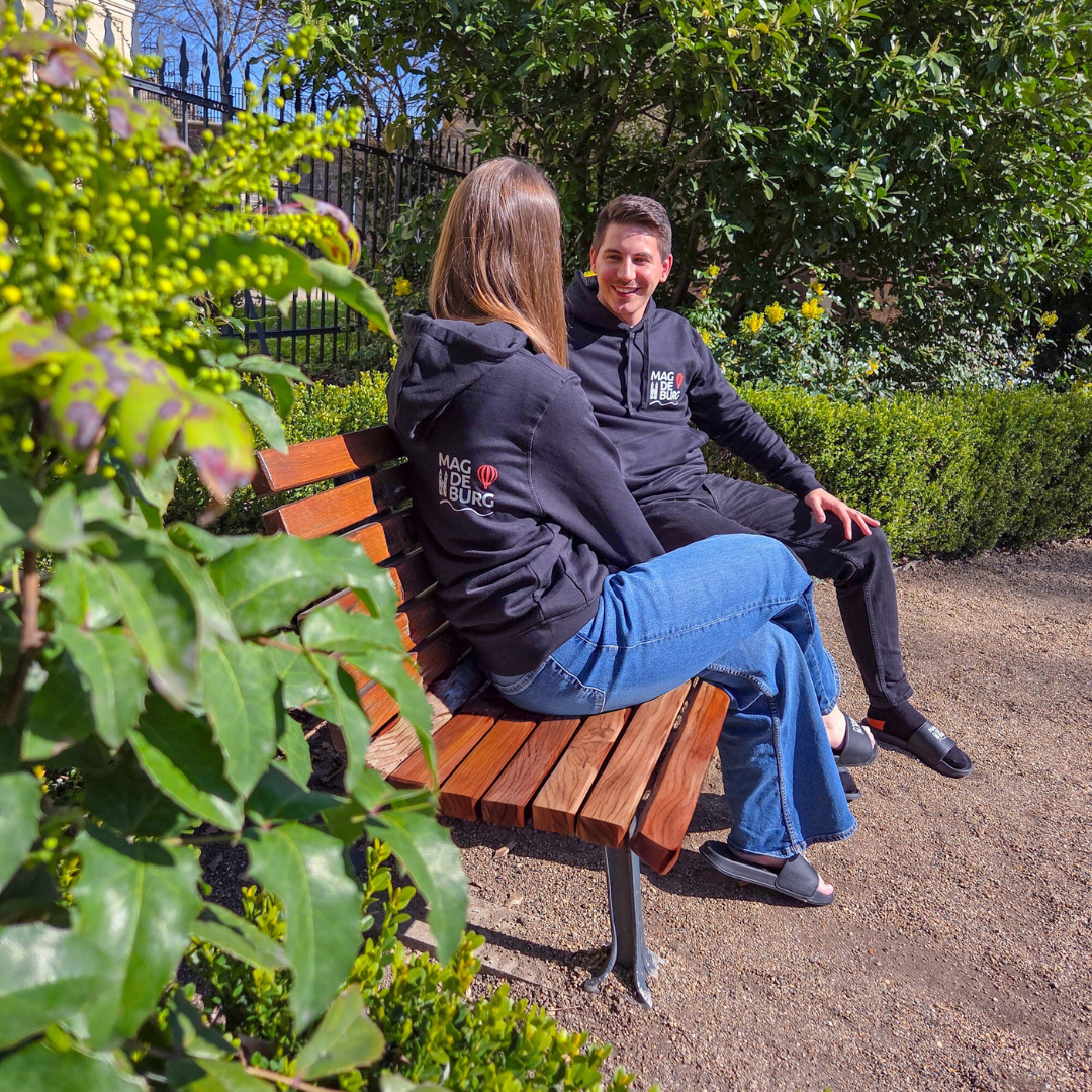 Zwei Personen sitzen auf einer Holzbank im Freien und tragen Magdeburg-Hoodies. Der Hoodie hat ein stilvolles Design mit einem auffälligen Logo. Die Szenerie ist umgeben von grünen Pflanzen, die einen entspannenden Park- oder Gartenlook vermitteln.