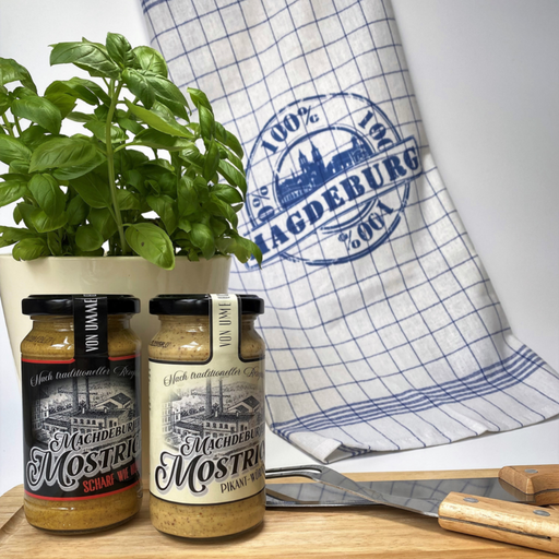 Zwei Gläser Machdeburjer Mostrich, pikant und würzig, auf einem Holzbrett, daneben ein Topf mit frischem Basilikum und ein Geschirrtuch mit Magdeburg-Logo, perfekt für eine stilvolle Küchenpräsentation – ideal für Liebhaber traditionellen deutschen Senfs.